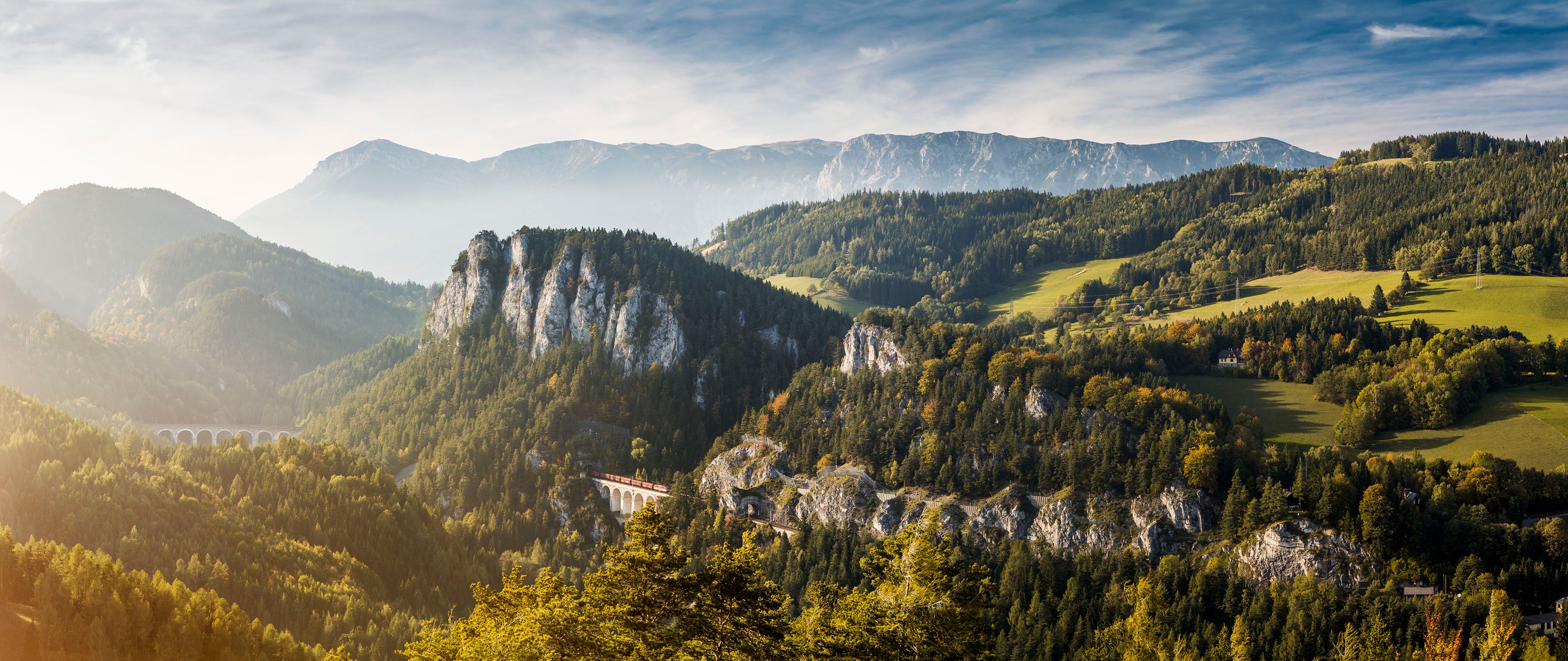 Panoramablick auf die Semmeringbahn in einer bergigen Landschaft mit Wäldern und einem Viadukt.