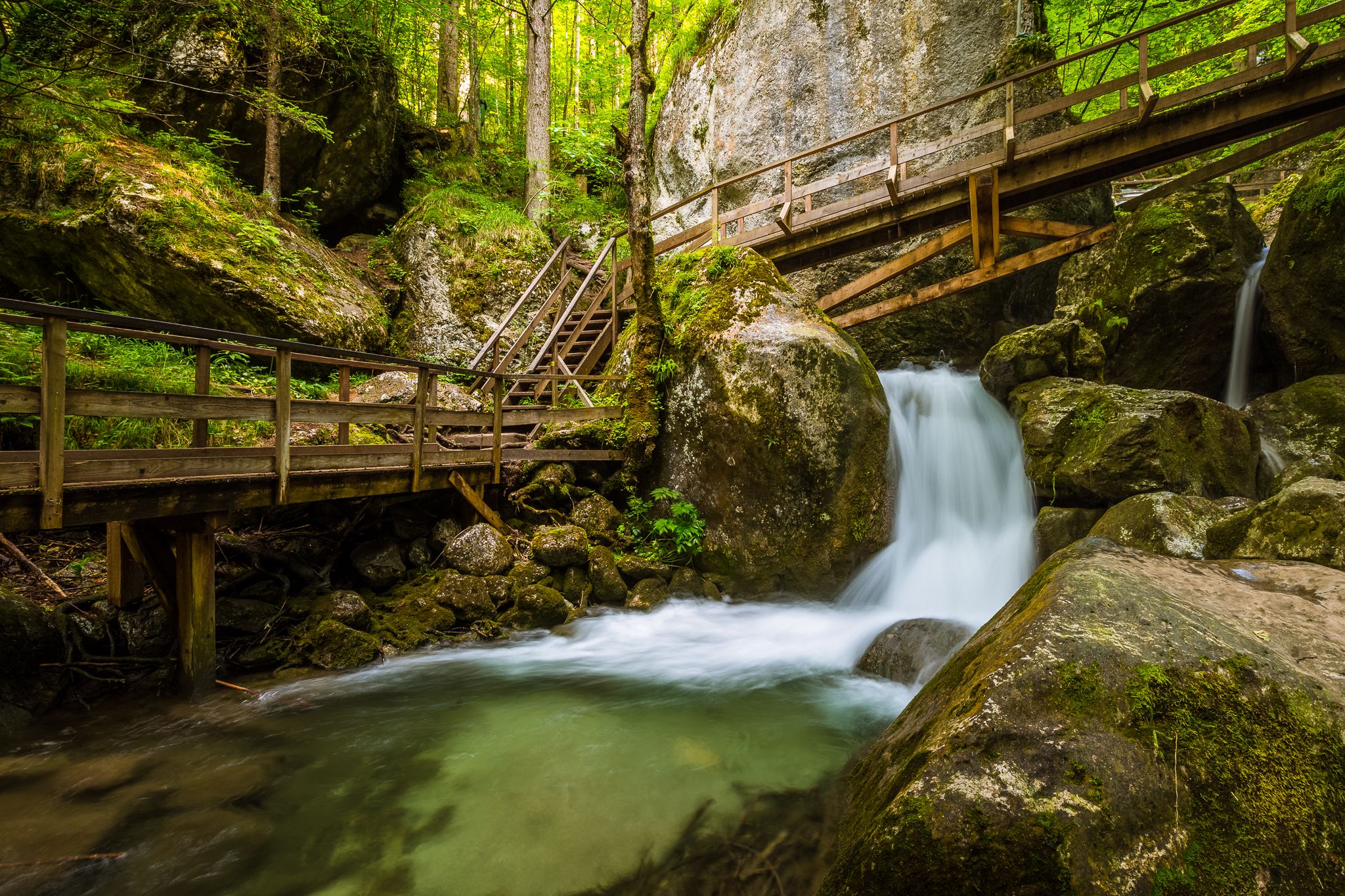 Wasserfall mit Holzbrücken und Treppen in einem bewaldeten Gebiet.