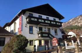 Ein traditionelles Gasthaus mit Holzverzierungen und einem Schild "Gasthaus Fam. Kamper" vor einem klaren blauen Himmel.