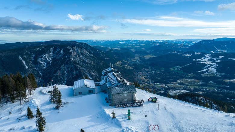 Luftaufnahme des Raxalm-Berggasthofs im Winter mit schneebedeckten Bergen und T&auml;lern im Hintergrund.