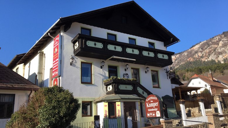 A traditional inn with wooden decorations and a sign "Gasthaus Fam. Kamper" in front of a clear blue sky.