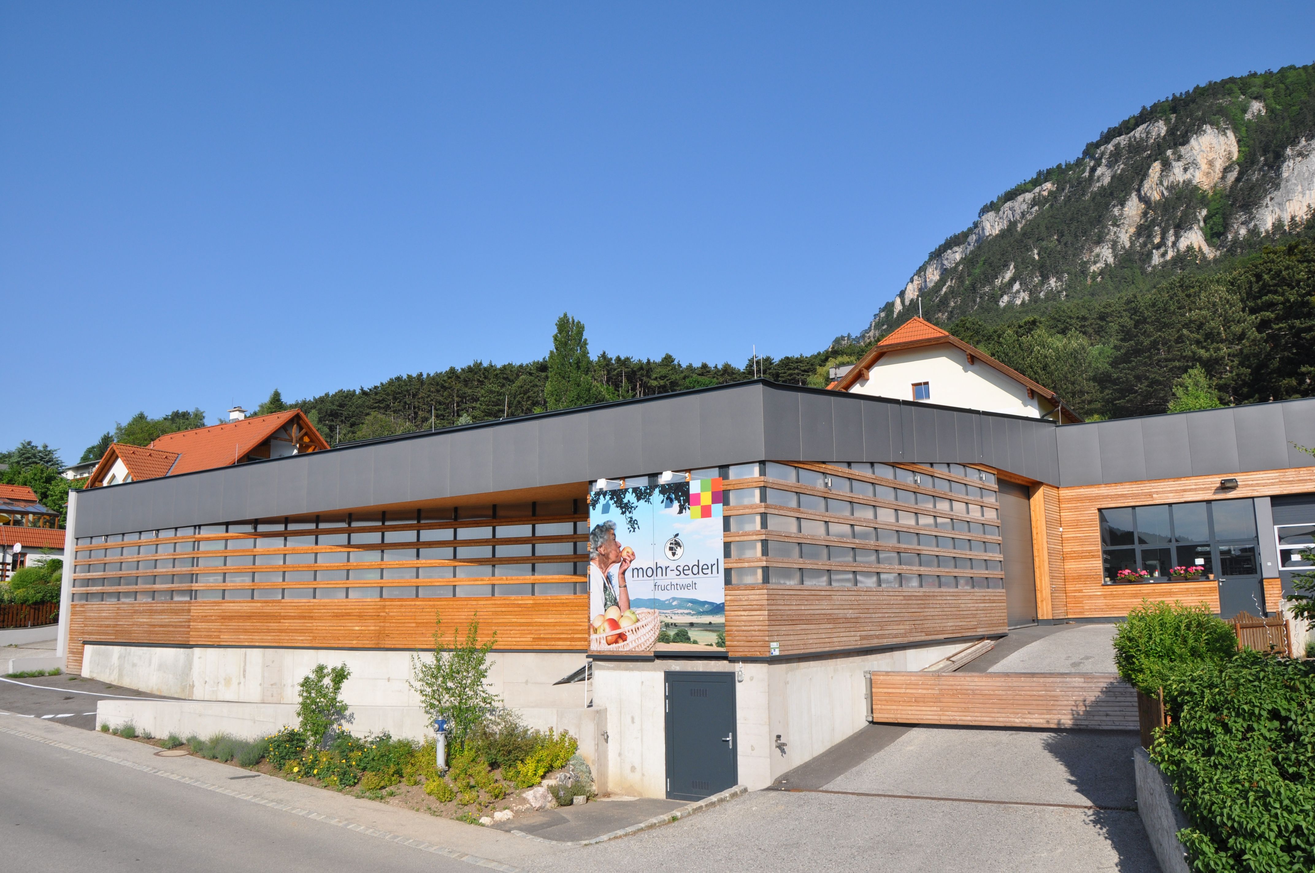 Modernes Gebäude mit Holzverkleidung und Werbeschild von Mohr-Sederl Fruchtwelt, umgeben von grüner Landschaft und Bergen.