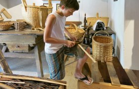 A boy works on a wood project in a museum, surrounded by baskets and traditional tools.