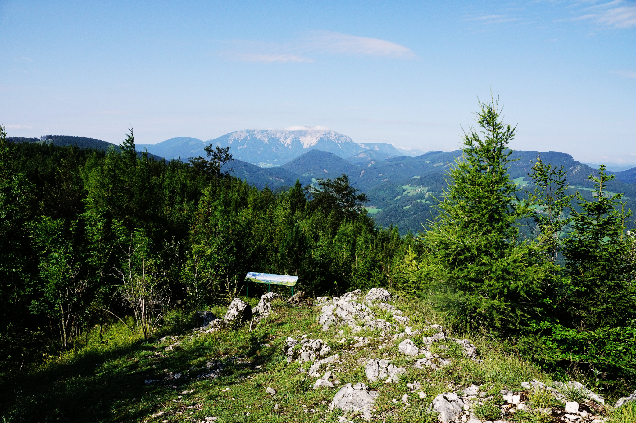 Blick vom Naturpark Hohe Wand auf den Schneeberg, mit bewaldeten Hügeln, Felsen und blauem Himmel.