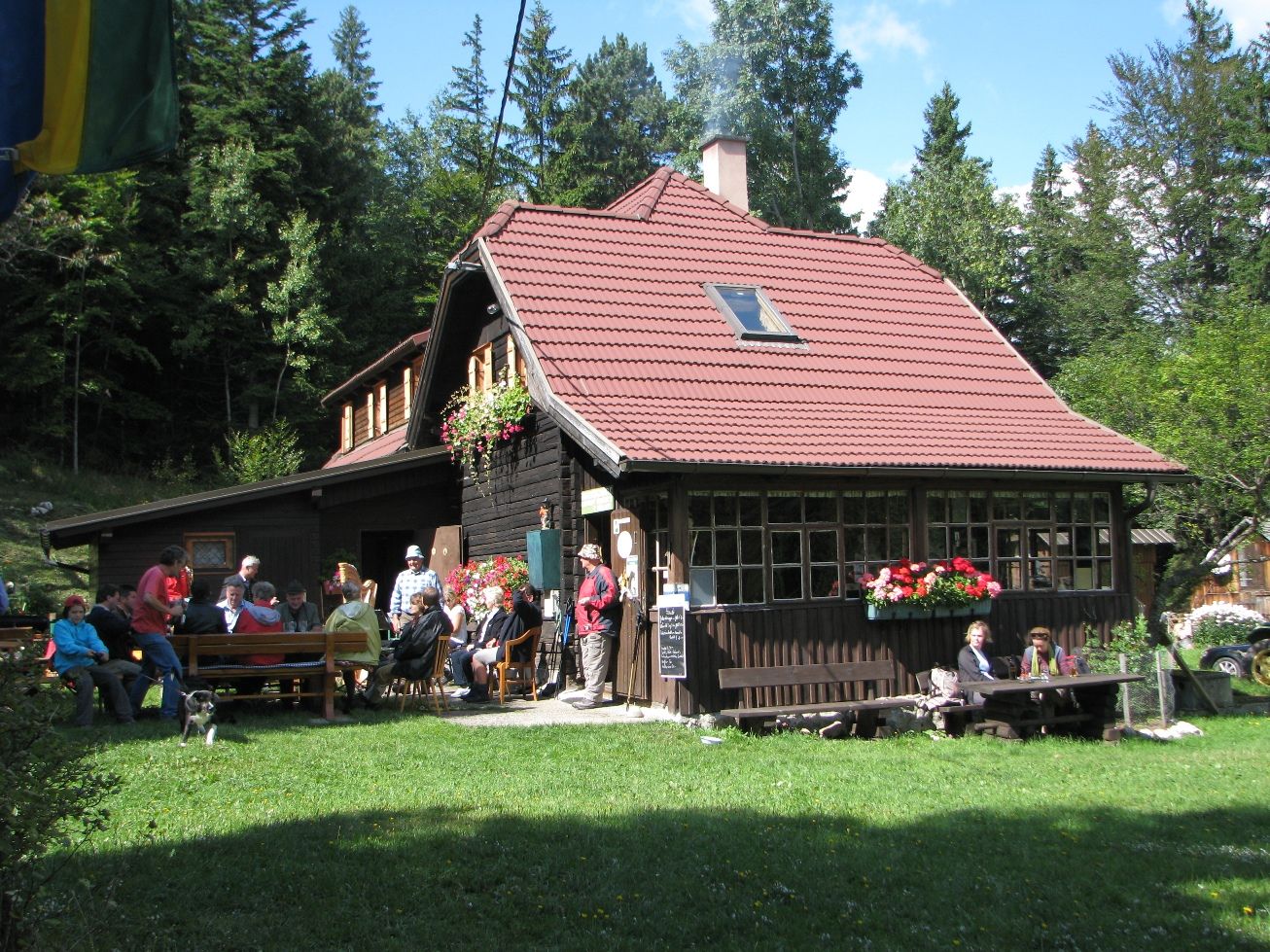 Eine Berghütte mit rotem Dach, umgeben von Wald, mit Menschen, die draußen sitzen.