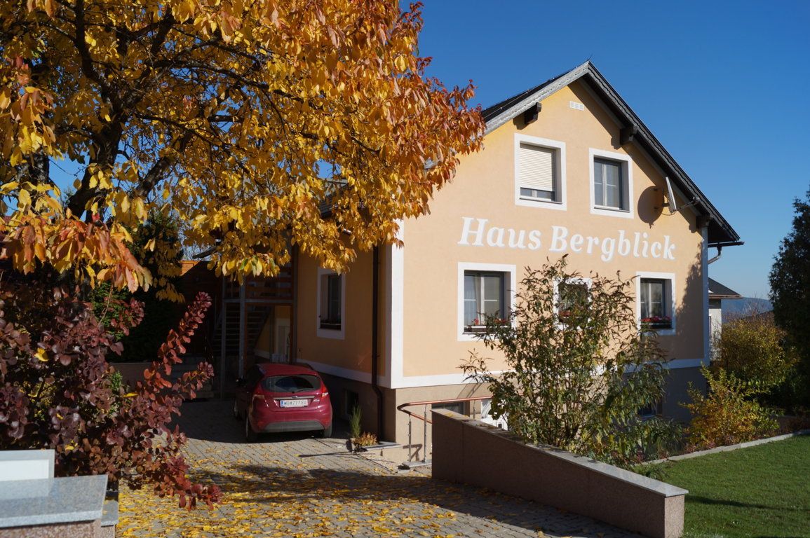 Egy sárga ház "Haus Bergblick" felirattal, körülötte őszi fák és egy parkoló autó az udvaron.