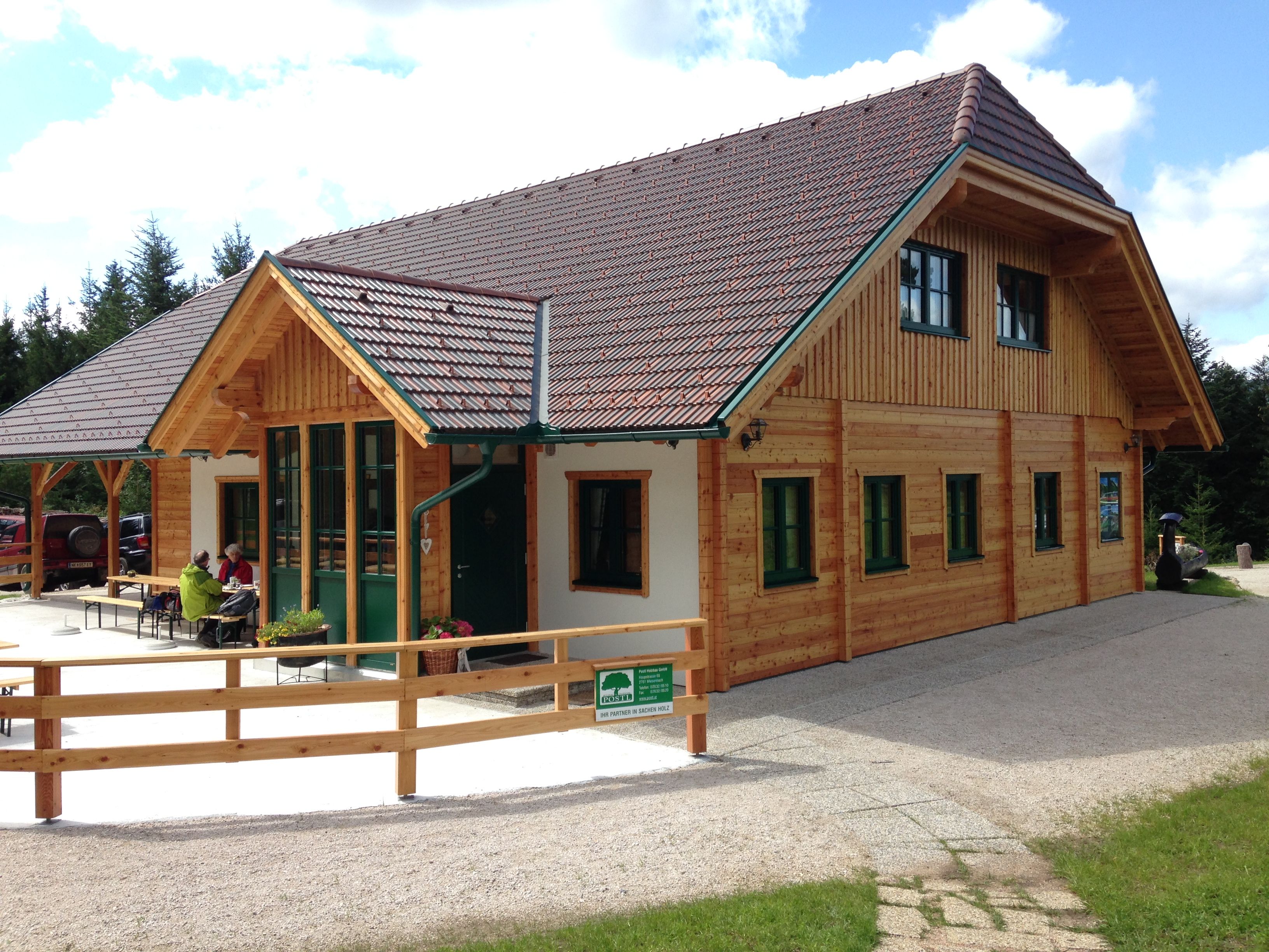 Eine Holzhütte mit grünem Dach und Fenstern, umgeben von Bäumen. Zwei Personen sitzen draußen an einem Tisch.