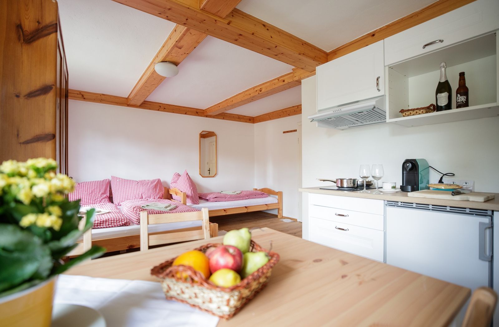 Gemütliche Ferienwohnung mit Holzmöbeln, einer kleinen Küche und einem Bett mit rot-weiß karierten Kissen. Ein Obstkorb steht auf dem Tisch.