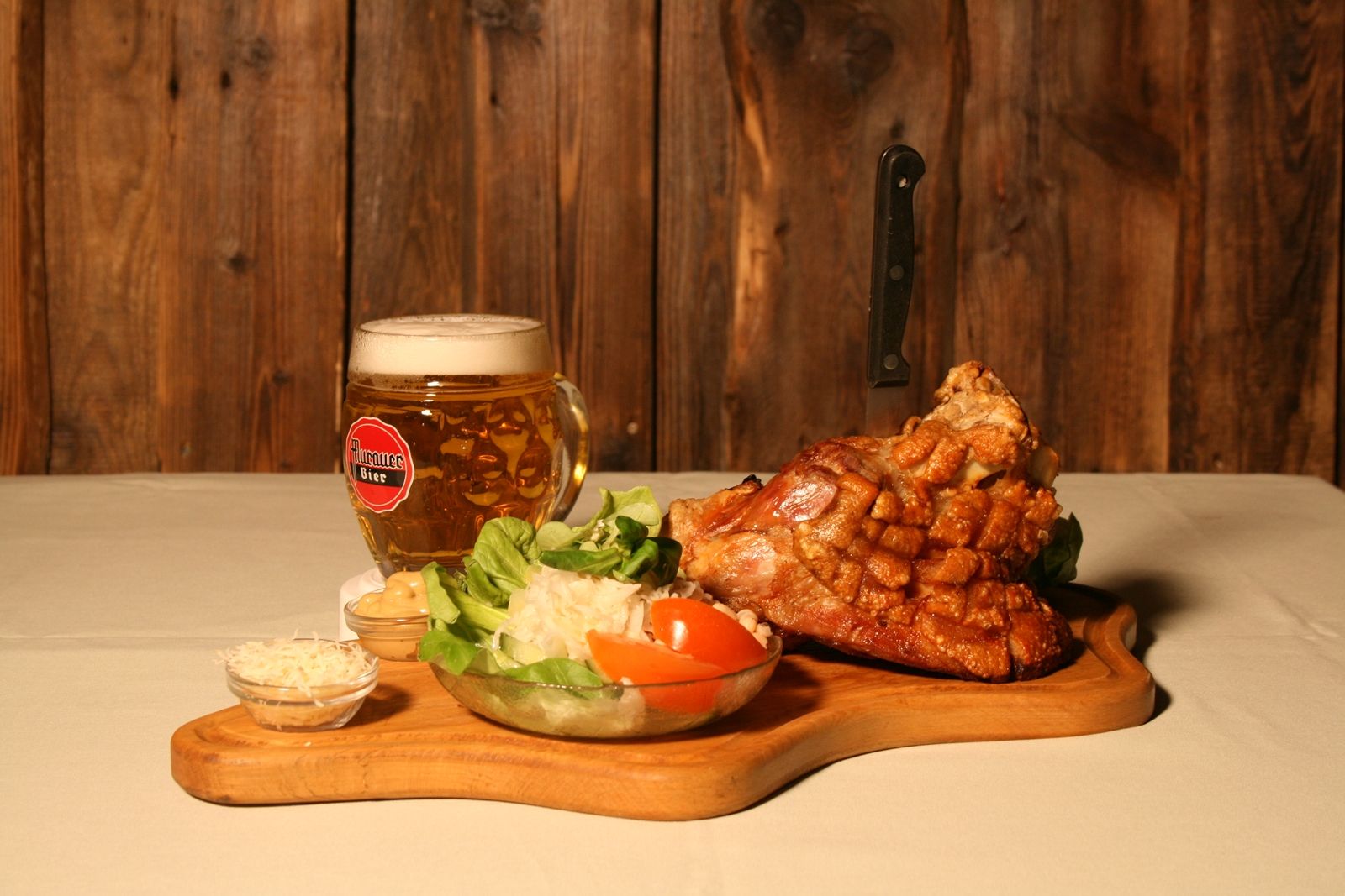 Ein Holzbrett mit Schweinshaxe, Salat und Bierkrug vor Holzwand.