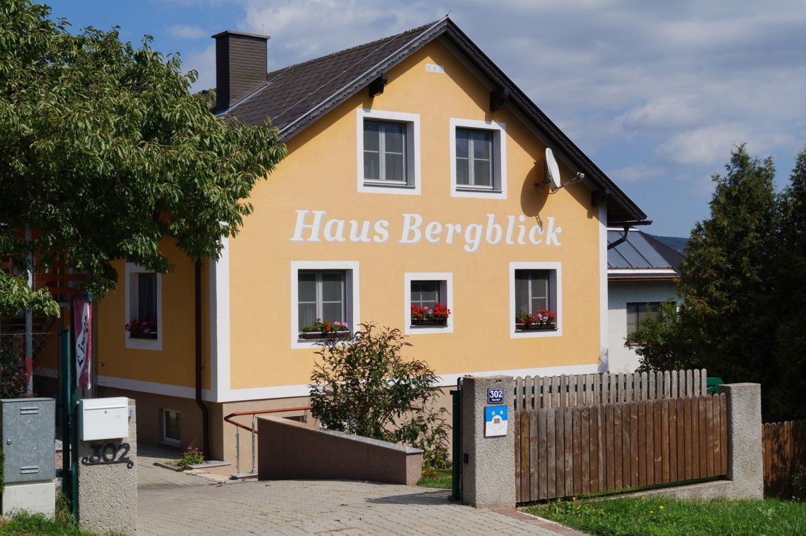 "Haus Bergblick" feliratú sárga ház, fákkal és fakerítéssel körülvéve.