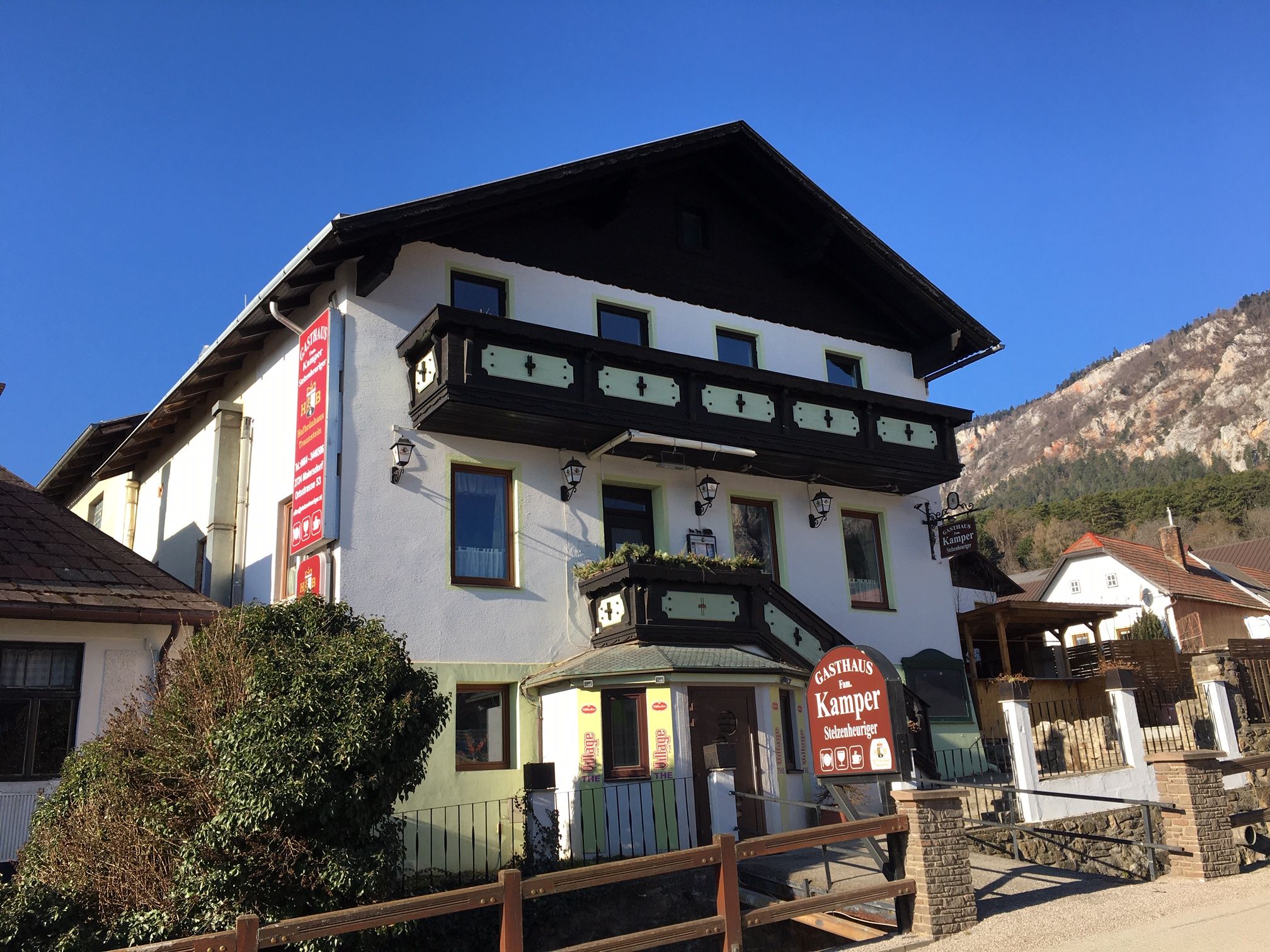 Ein traditionelles Gasthaus mit Holzverzierungen und einem Schild "Gasthaus Fam. Kamper" vor einem klaren blauen Himmel.