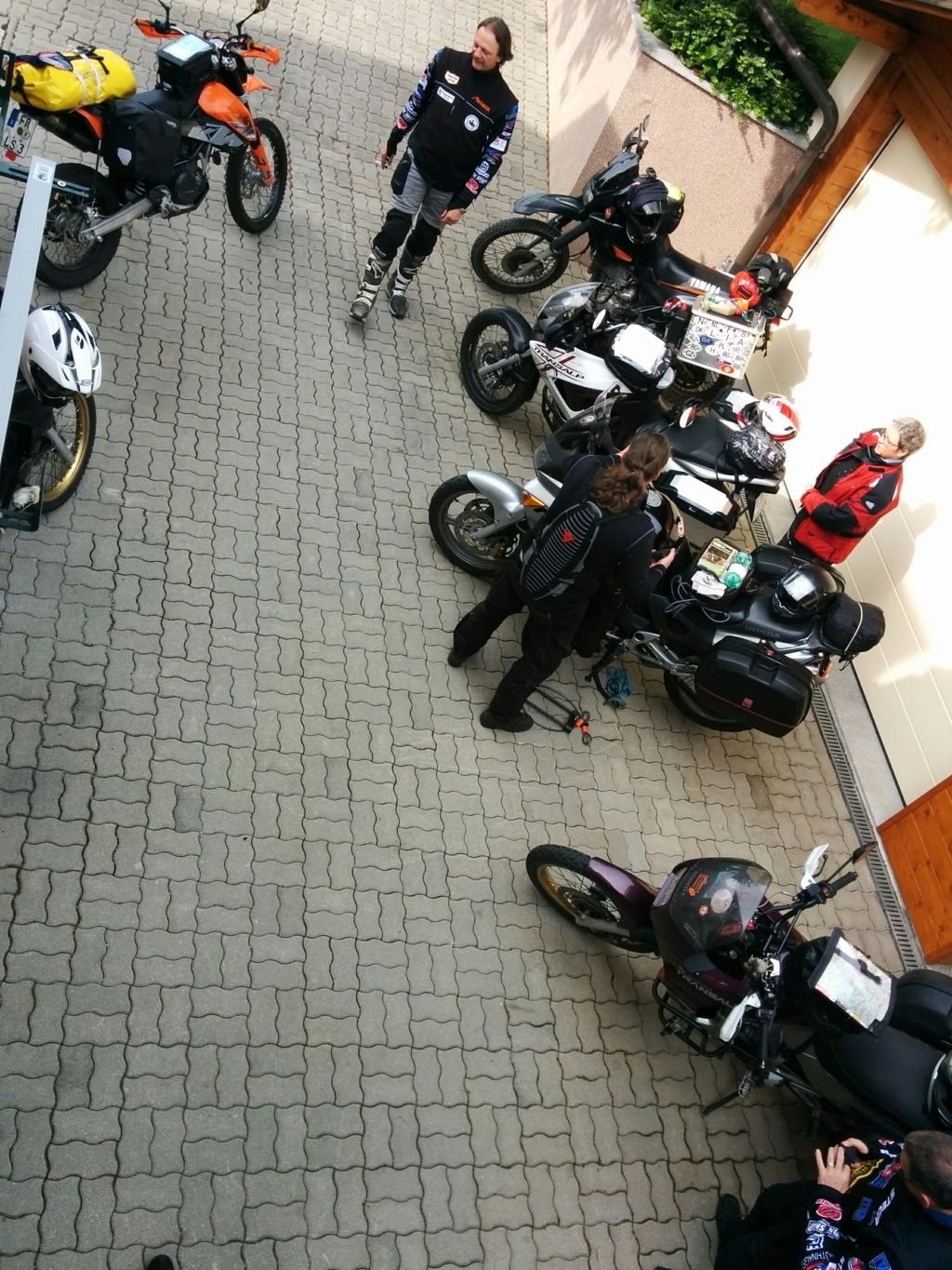Több motorkerékpár és emberek egy kikövezett téren, egy épület előtt.