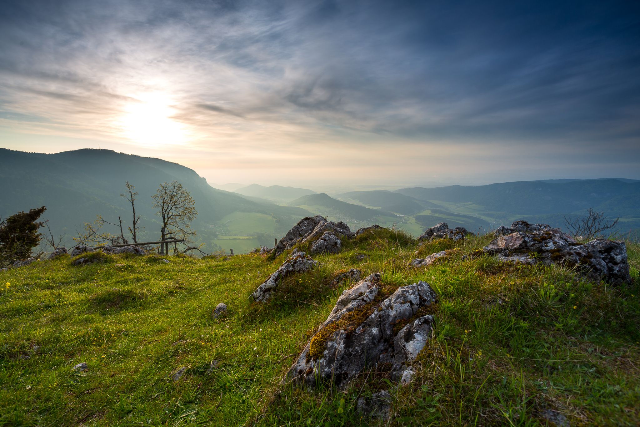 Panoramablick von einem Hügel mit Felsen und Gras im Vordergrund, Bergen und einem bewölkten Himmel im Hintergrund.