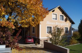 A yellow house with the inscription 'Haus Bergblick', surrounded by autumnal trees and a parked car in the courtyard.
