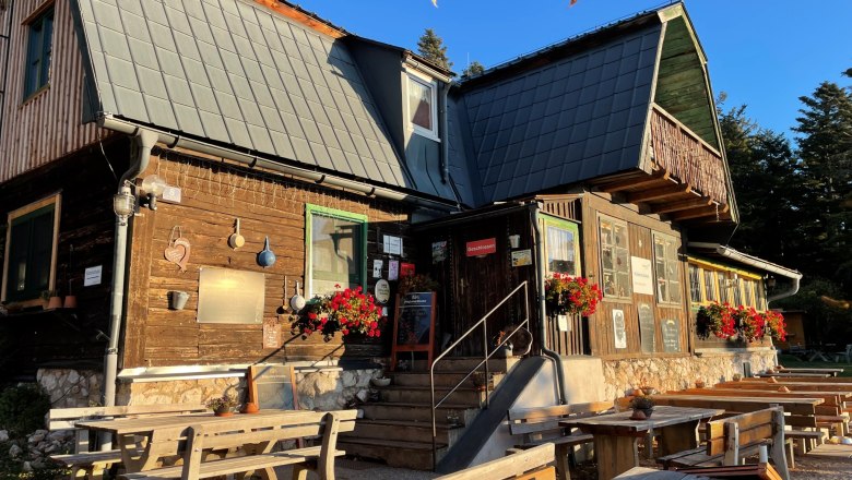 Ein rustikales Holzhaus mit Blumen und Sitzb&auml;nken im Freien bei Sonnenschein.