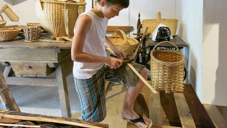 A boy works on a wood project in a museum, surrounded by baskets and traditional tools.