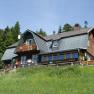 A traditional mountain house with a wooden fa&ccedil;ade and shingle roof, surrounded by trees and meadows.
