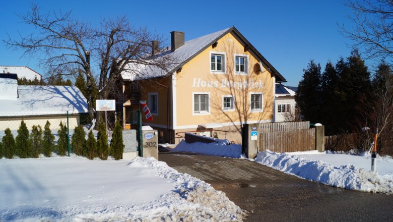 Ein gelbes Haus mit der Aufschrift 'Haus Bergblick' im Schnee, umgeben von B&auml;umen und einem blauen Himmel.