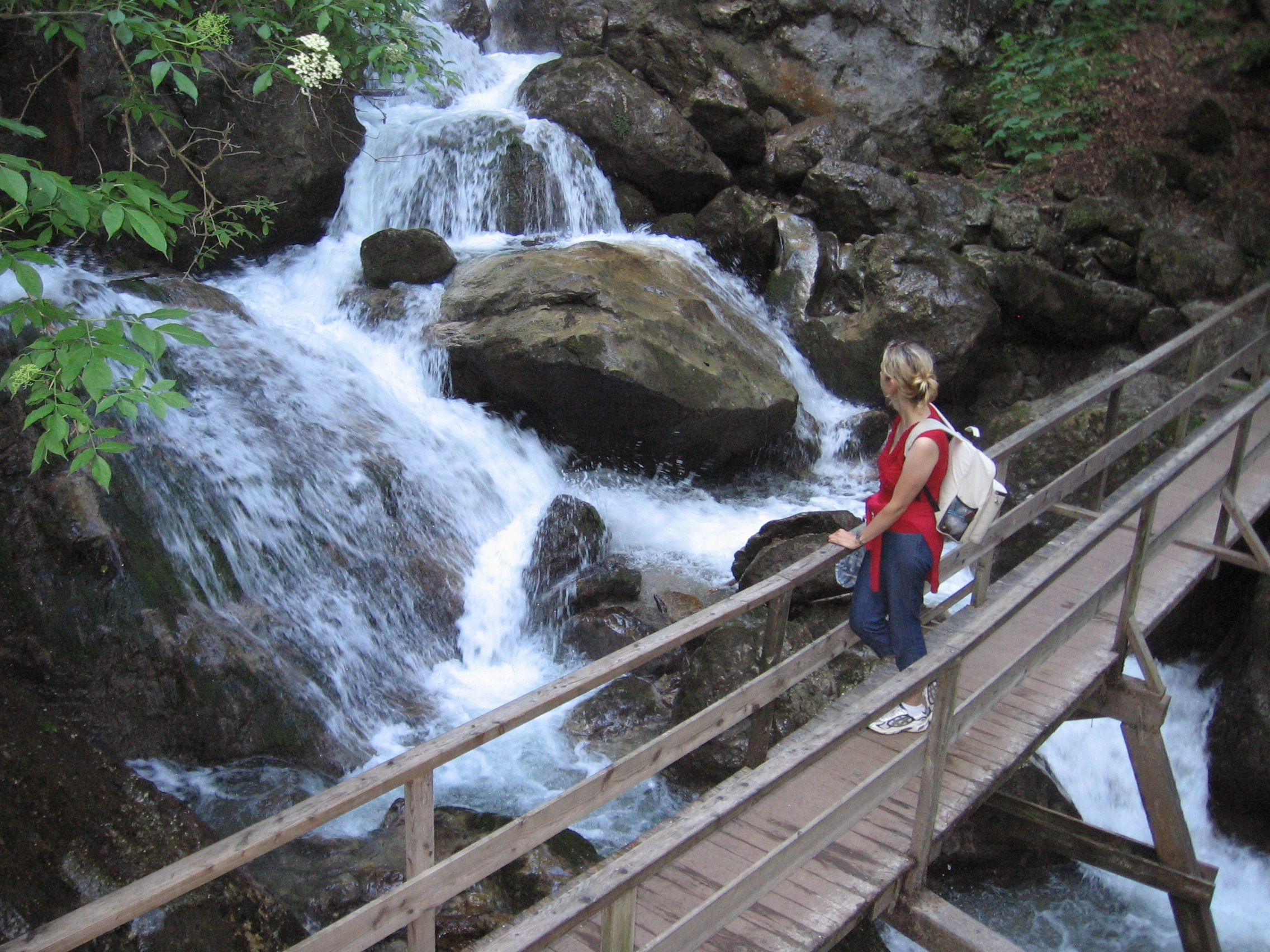 Person auf Holzbrücke bei Wasserfall in bewaldeter Umgebung.