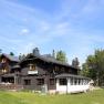 A mountain hut with dark wood and white windows, surrounded by trees and meadows, under a blue sky.