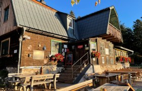 Ein rustikales Holzhaus mit Blumen und Sitzb&auml;nken im Freien bei Sonnenschein.