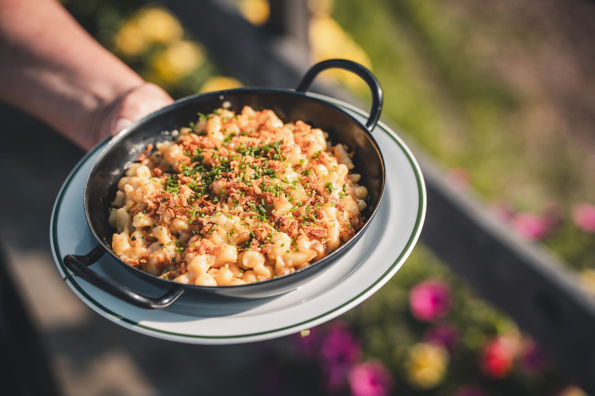 Eine Pfanne voll Käsespätzle, appetitlich serviert mit Zwiebel und Schnittlauf in einer rustikalen Pfannne und Blick ins Freie.