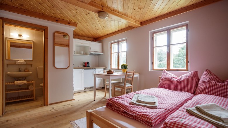 Gem&uuml;tliche Ferienwohnung mit Holzboden, Doppelbett, kleiner K&uuml;che und Esstisch. Badezimmer im Hintergrund.