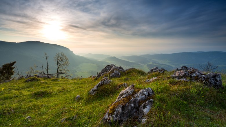 Panoramablick von einem Hügel mit Felsen und Gras im Vordergrund, Bergen und einem bewölkten Himmel im Hintergrund.