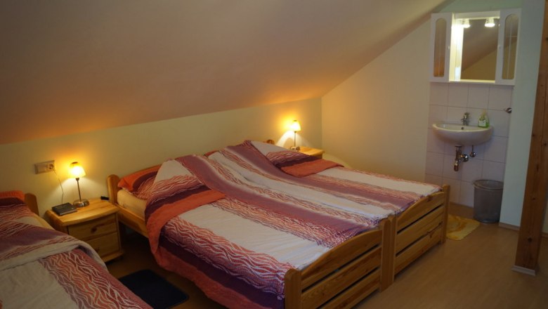 A room with three beds, bedside tables and a washbasin under a sloping roof.
