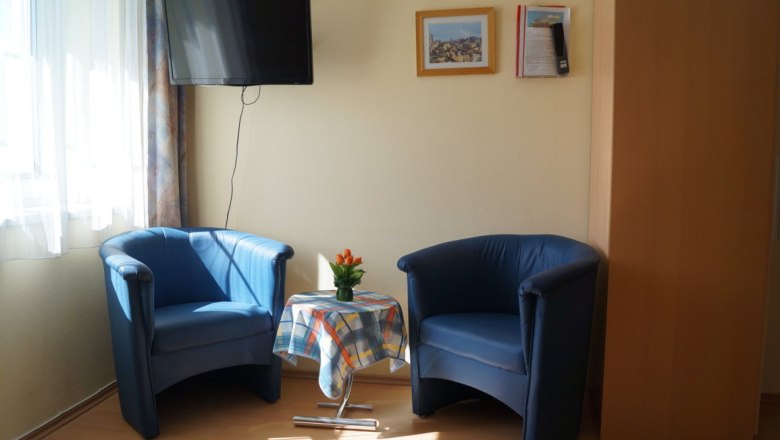 Zwei blaue Sessel mit einem kleinen Tisch dazwischen, darauf eine Vase mit Blumen. Ein Fernseher h&auml;ngt an der Wand, daneben zwei Bilder.