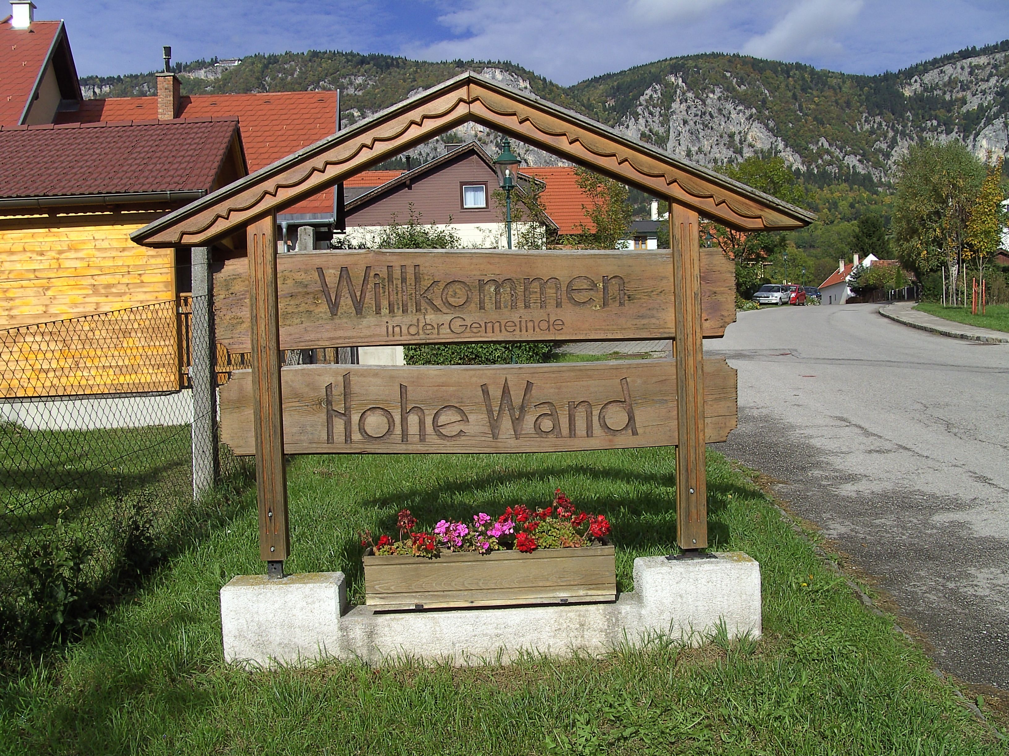 Fából készült tábla "Üdvözöljük Hohe Wand településen" felirattal egy hegyvidéki táj előtt.