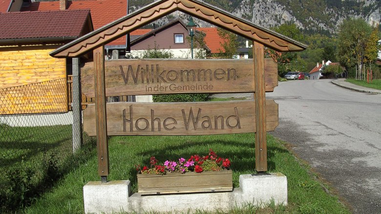 Wooden sign with the inscription 'Welcome to the municipality of Hohe Wand' in front of a mountain landscape.