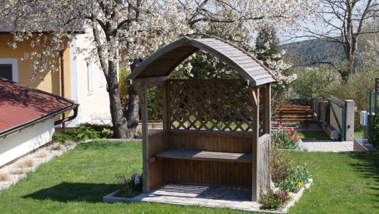 Holzbank mit Überdachung im Vorgarten, umgeben von blühenden Bäumen und Blumen.