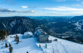 Luftaufnahme des Raxalm-Berggasthofs im Winter mit schneebedeckten Bergen und Tälern im Hintergrund.