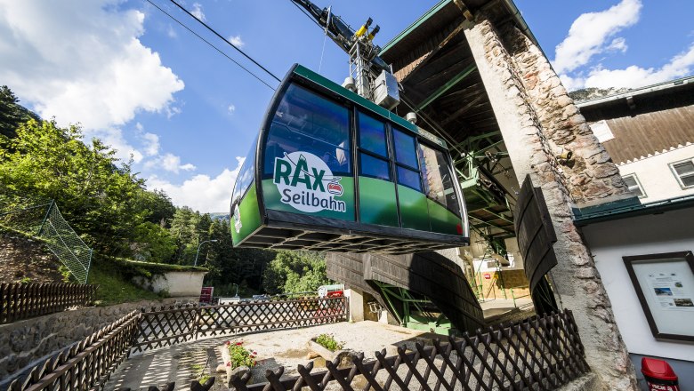 Rax-Seilbahn bei der Talstation, umgeben von B&auml;umen und blauem Himmel.