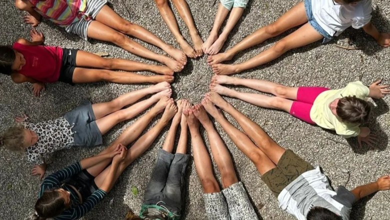 Children sit in a circle on gravel, legs stretched out towards the center.