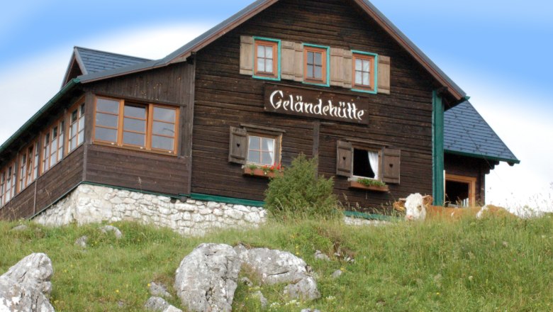 Eine Berghütte mit der Aufschrift 'Geländehütte' auf einer Wiese mit Felsen und einer Kuh im Vordergrund.