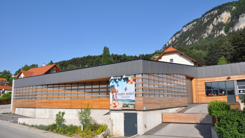 Modern building with wooden cladding and advertising sign for Mohr-Sederl Fruchtwelt, surrounded by green countryside and mountains.