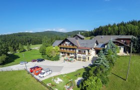 Alpengasthof Postl in einer grünen, bewaldeten Landschaft mit mehreren geparkten Autos und einem Spielplatz im Vordergrund.