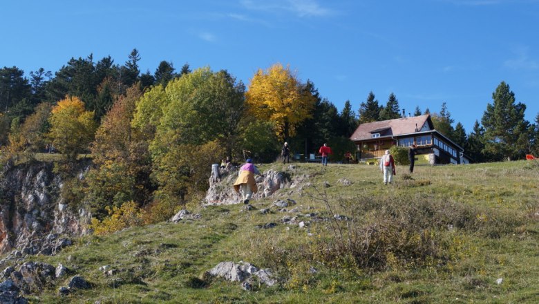 Menschen wandern auf einem grasbewachsenen H&uuml;gel mit einer Bergh&uuml;tte im Hintergrund.