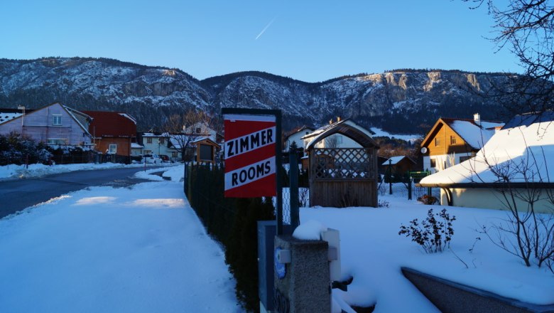 Winter landscape with snow-covered houses and mountains in the background. A sign with the inscription 'Zimmer Rooms' is visible.