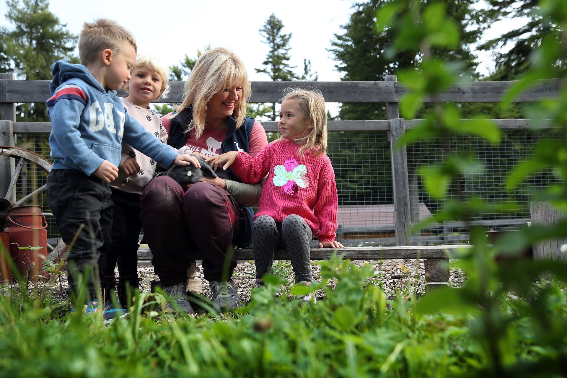 Eine Naturpark-Mitarbeiterin sitzt mit einem Kaninchen am Schoß im Streichelzoo des Naturpark Hohe Wand und wird von 3 Kindern umgeben, welche das Kaninchen streicheln. 