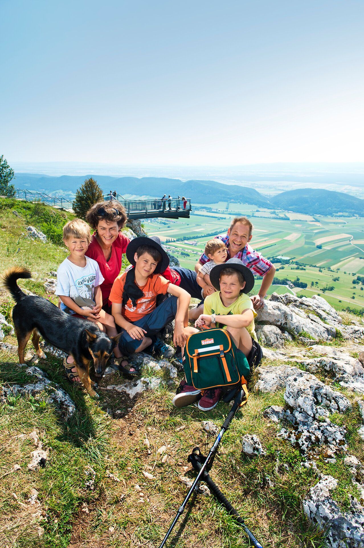 Eine fröhliche Familie genießt die atemberaubende Aussicht vom Skywalk, umgeben von der majestätischen Berglandschaft. Die Kinder spielen mit ihrem Hund, während die Eltern die frische Bergluft und die malerische Kulisse bewundern. Hier wird der Sommer in den Bergen zu einem unvergesslichen Erlebnis.