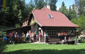 A mountain hut with a red roof, surrounded by forest, with people sitting outside.