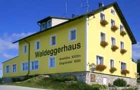 Gelbes Gebäude mit der Aufschrift 'Waldeggerhaus', umgeben von Blumen.