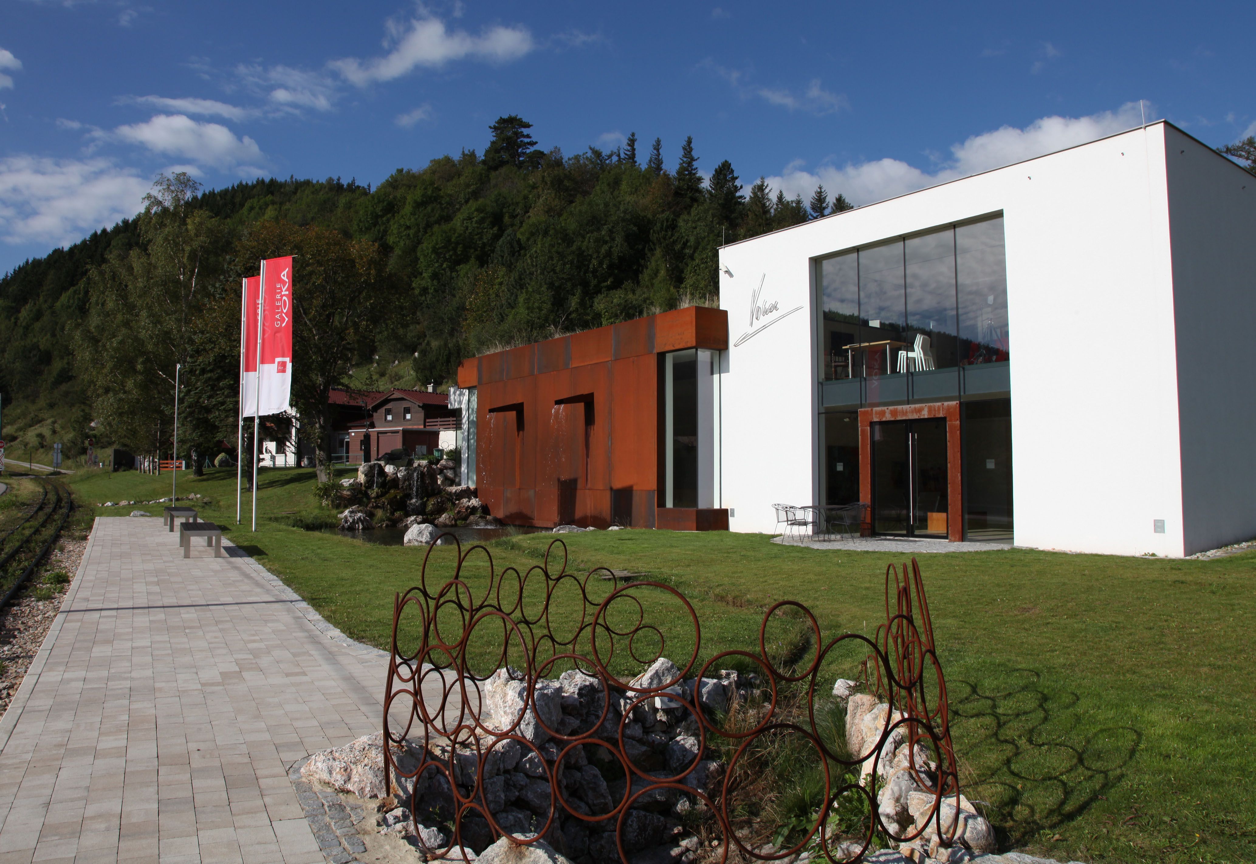 Modern gallery with glass façade and rusty metal sculptures in a green setting.