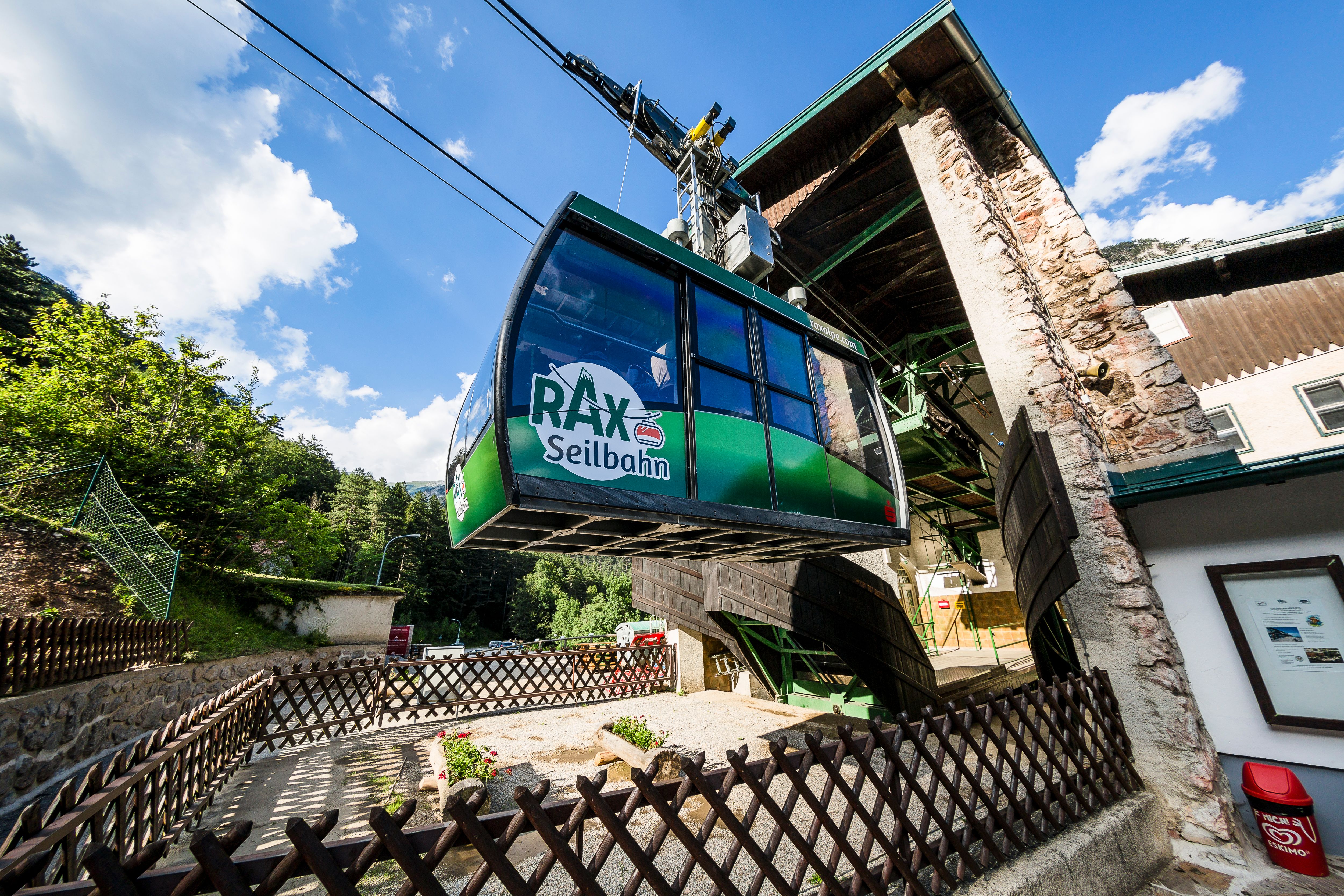 Rax-Seilbahn bei der Talstation, umgeben von Bäumen und blauem Himmel.