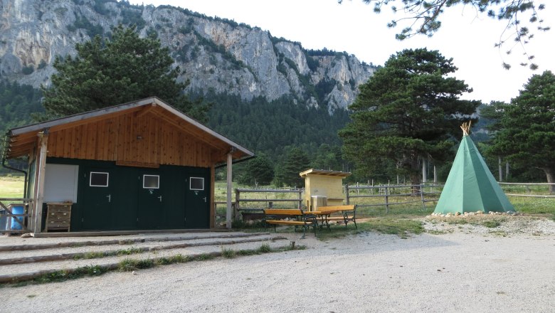 Ein Zeltplatz mit einem grünen Tipi, einer Holzhütte und Bänken vor einer Felswand und Bäumen.