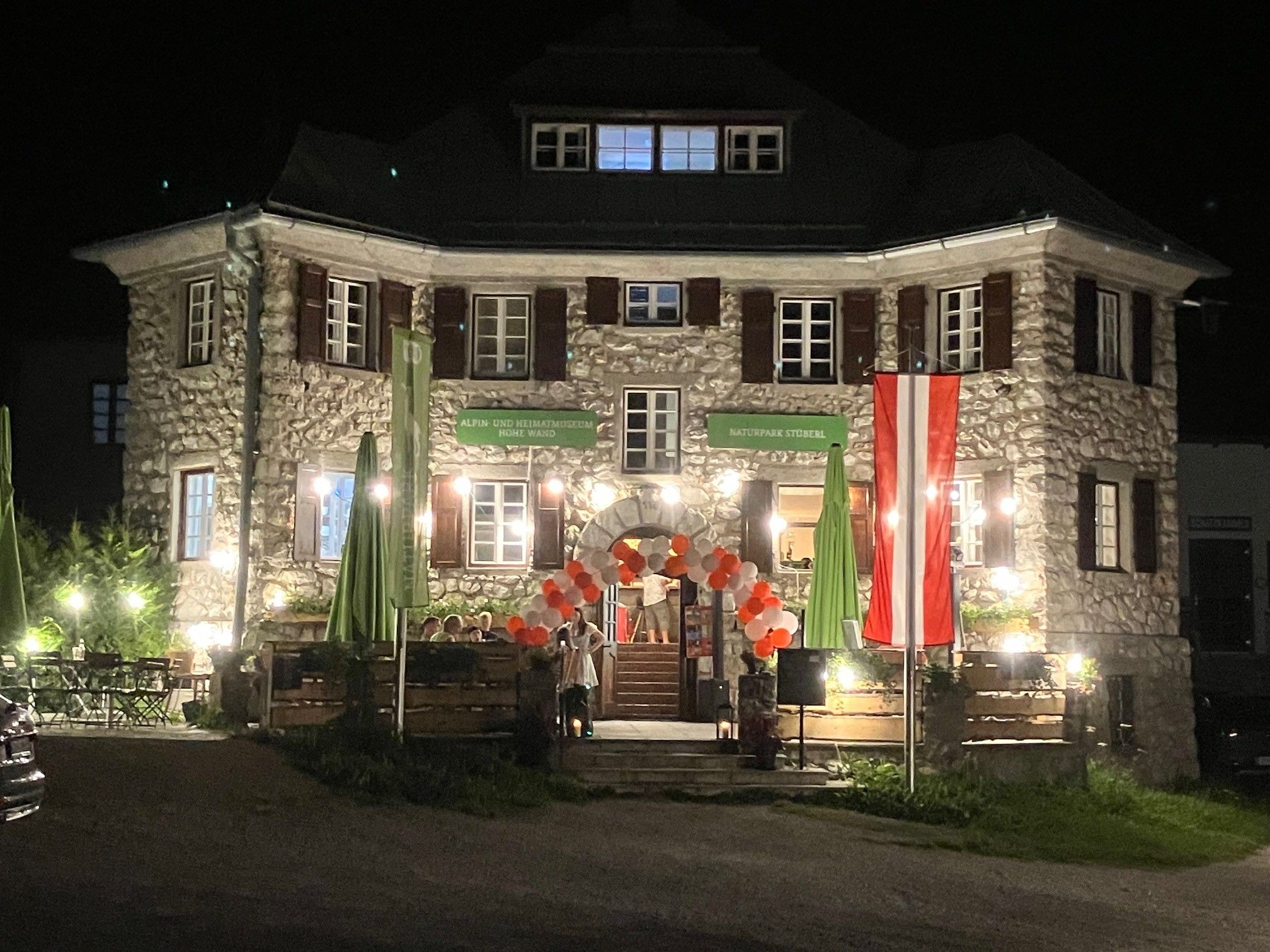 Ein beleuchtetes Steingebäude bei Nacht mit österreichischer Flagge und Luftballons am Eingang.