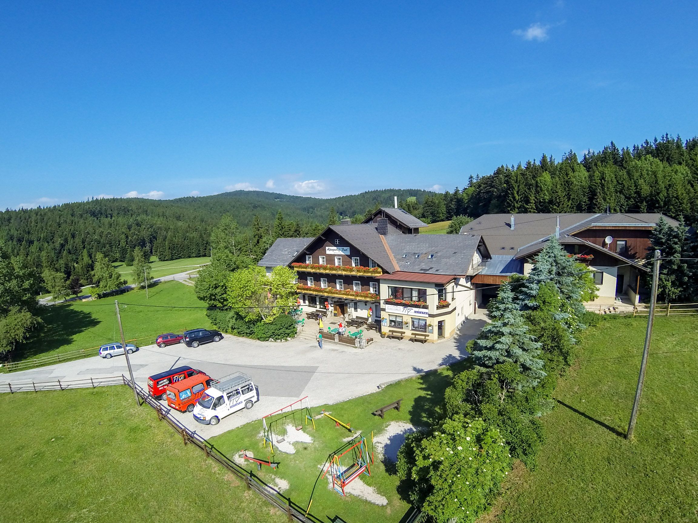 Alpengasthof Postl in einer grünen, bewaldeten Landschaft mit mehreren geparkten Autos und einem Spielplatz im Vordergrund.